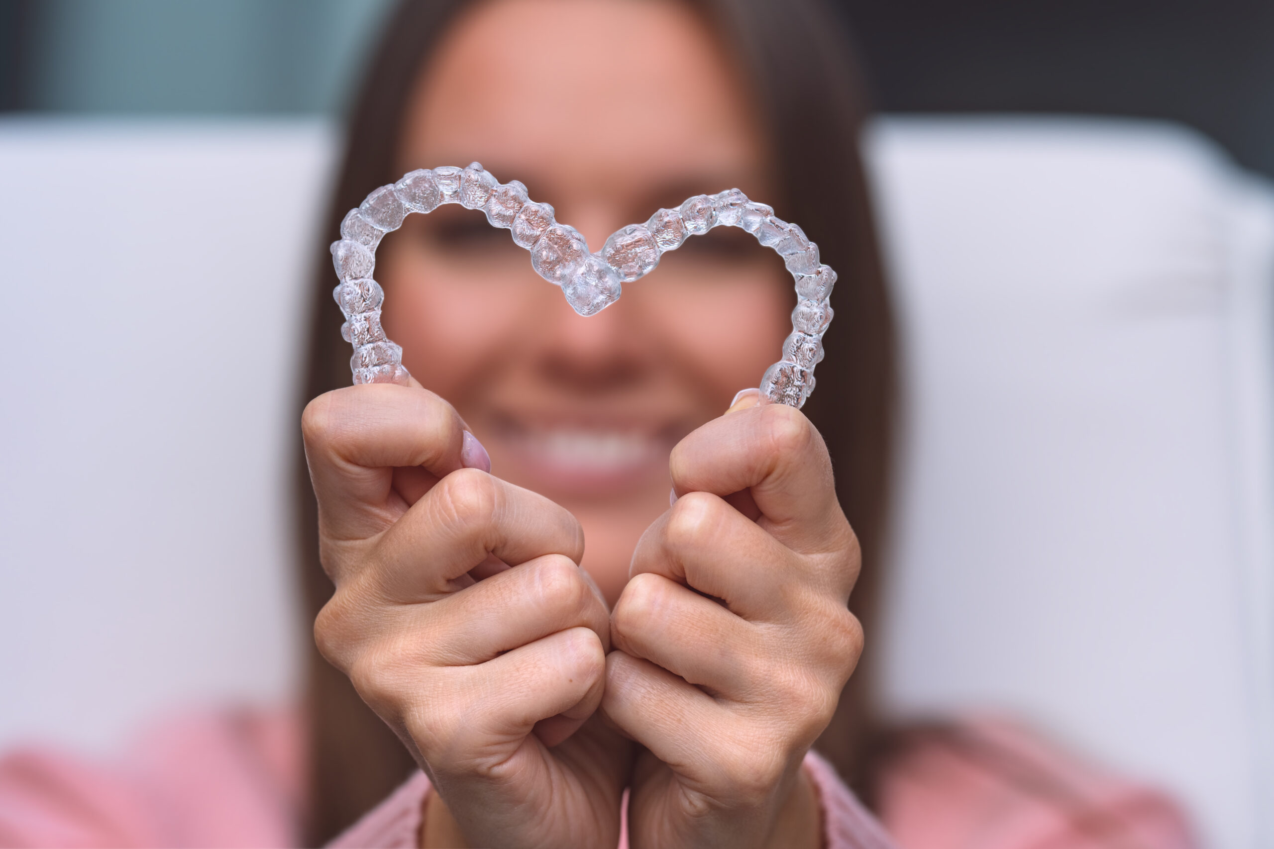 Woman with dental invisible invisalign braces or silicone trainer. Aligners treatment Smiling woman holding Invisalign clear aligners in a heart shape, invisible braces available at The Center for Complete Healthy Dentistry in Wilton, CT
