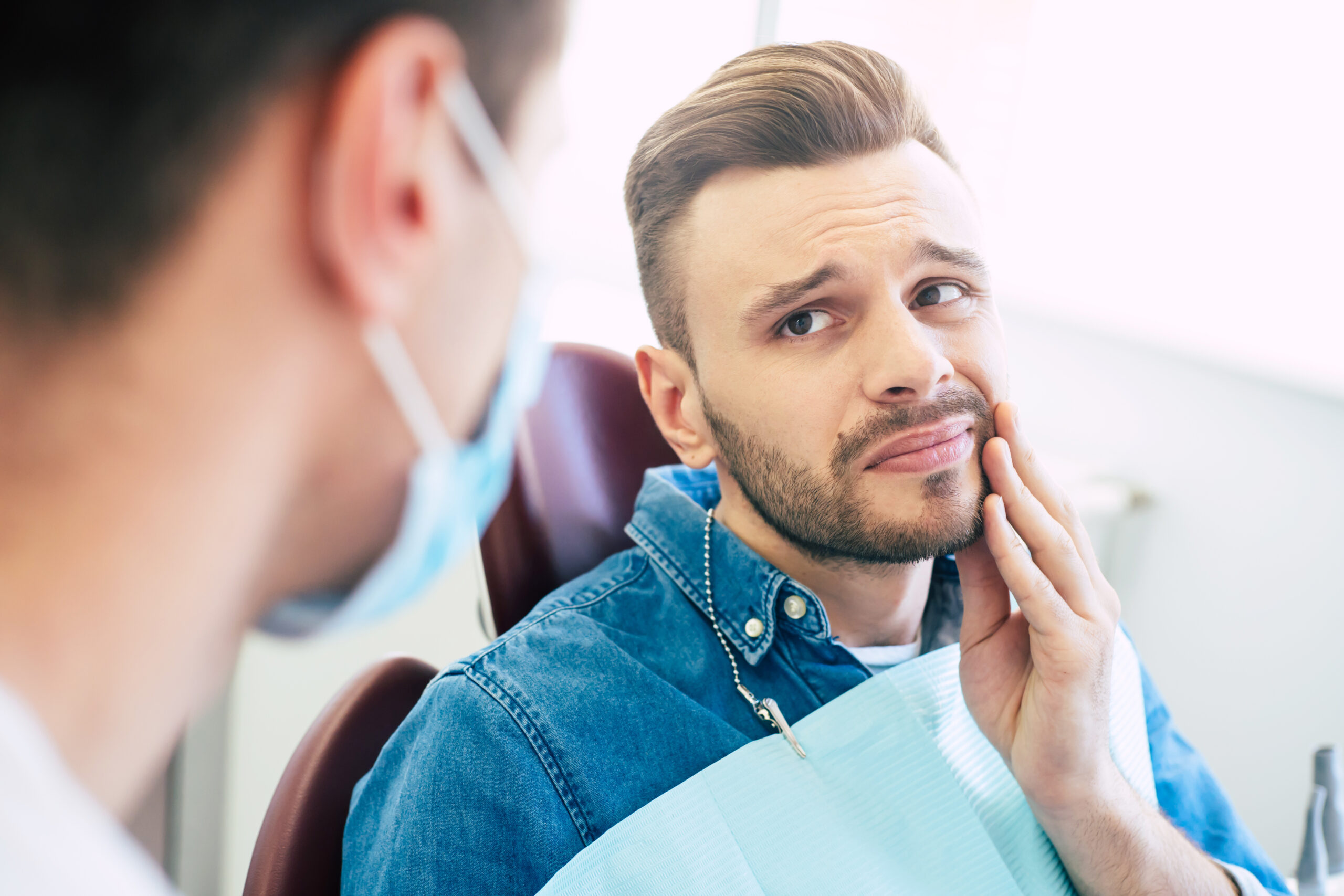 Symptoms of gums pain. A man with a worried face is holding his hand on his cheek because of irritating pain in front of a dentist who is going to give a patient a treatment. Patient experiencing jaw pain from TMJ/TMD discussing symptoms with dentist at The Center for Complete Healthy Dentistry in Wilton, CT